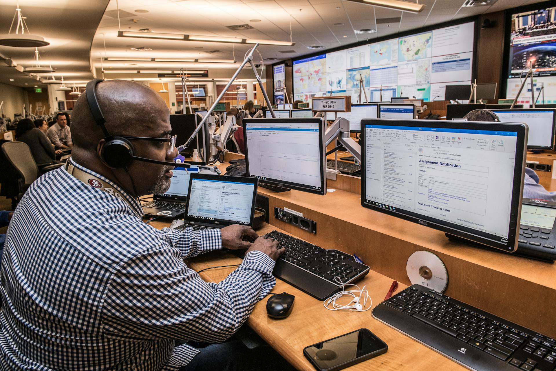 Man in Black And White Checkered Dress Shirt Using Computer in computing command center with other personel
